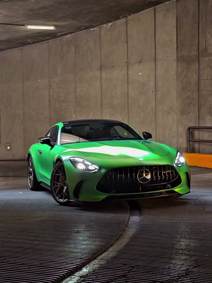 Mercedes-AMG GT verde satinado, frontal completo y centrado, solo en el cuadro, en un estacionamiento subterráneo con muro de concreto y franjas amarillas de fondo. Se ven la parri