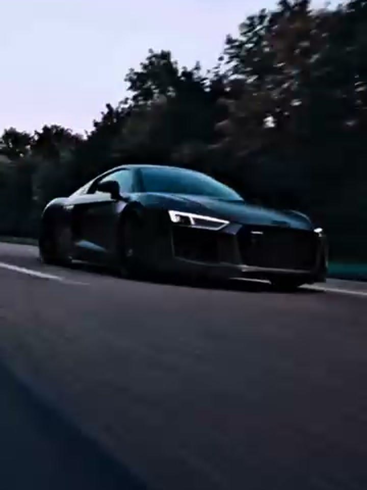 Audi R8 en negro satinado (PPF stealth), tres cuartos frontal completo, estacionado en el taller sobre piso con marcas blancas de posicionamiento; faros LED encendidos, parrilla de