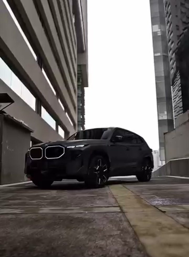 BMW XM completa en tres cuartos frontal, parada en un callejón entre torres de Santa Fe, piso mojado, luz de día nublado. Carrocería en acabado satinado muy oscuro (lee negro, no g