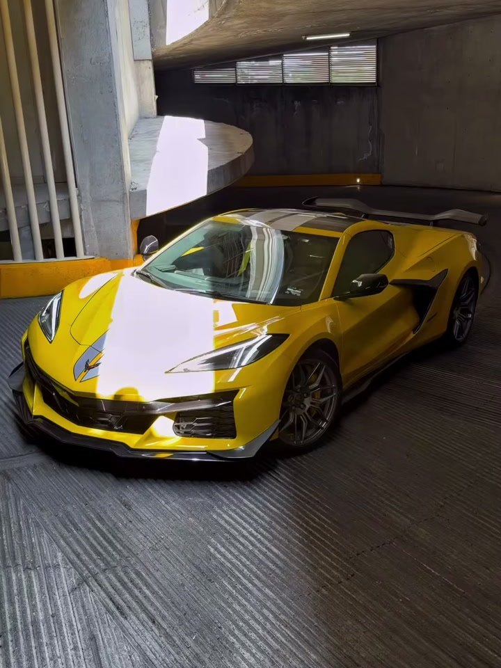 Corvette C8 Z06 amarillo completo, tres cuartos trasero en contrapicado, bajo sol pleno frente a la entrada de un estacionamiento: alerón alto, calaveras, difusor negro y los cuatr