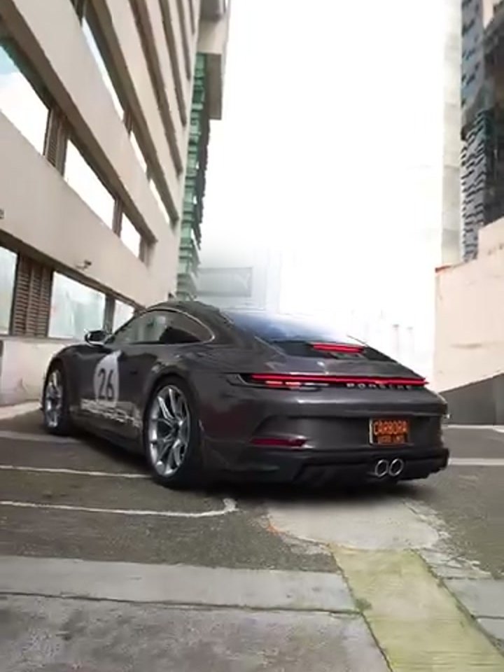 Porsche 911 (992) en gris grafito metálico, tres cuartos frontal completo desde ángulo bajo, en el estacionamiento de azotea entre torres. Se leen el rondel blanco con el «26» en l